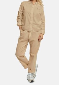 Camicia in lino beige con colletto e bottoni, abbinata a pantaloni in stile jogger coordinati. Tessuto leggero e vita con cordoncino, completata da sneakers bianche.