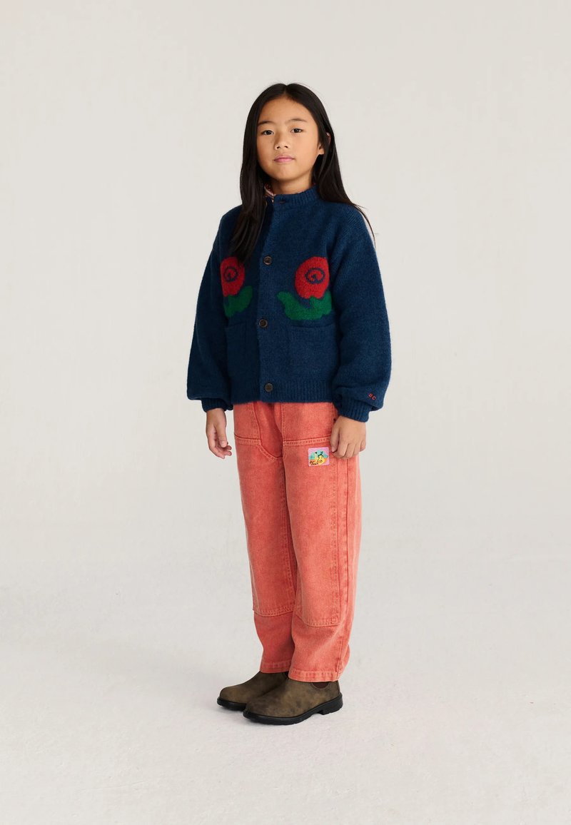 Jeune fille portant un cardigan bleu marine avec des motifs floraux rouges, un pantalon corail à jambes larges avec un patch, et des bottes marron, debout devant un fond uni.