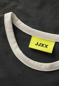 Černý límec trička s kontrastním bílým lemem; uvnitř výstřihu je viditelná žlutá visačka s potiskem "JJXX" černou barvou.