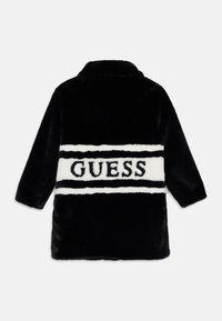 Čierny kožuch z falošnej kožušiny s bielym vodorovným pruhom, na ktorom je čierny text "GUESS". Má zárezový golier a dlhé rukávy. Hladká textúra.
