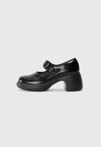 Black leather Mary Jane shoe with a round toe, adjustable buckle strap, and a thick, textured heel, featuring a smooth surface and minimal design.