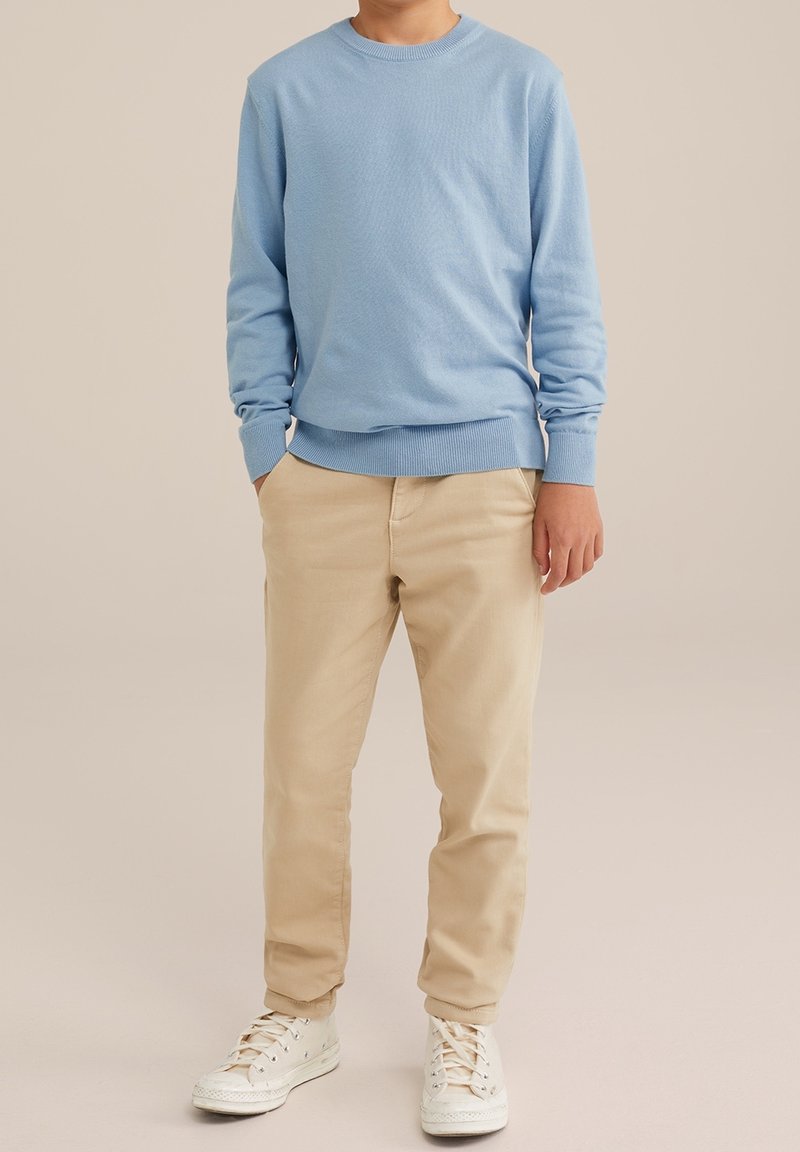 Enfant portant un pull bleu clair, un pantalon beige et des baskets blanches, debout avec les mains partiellement dans les poches sur un fond neutre.