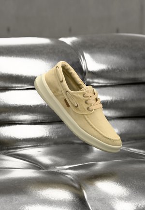 Chaussure bateau en toile beige avec semelle et lacets crème flottant au-dessus d'une surface en cuir argenté brillant capitonné.