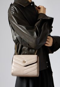 Borsa a tracolla trapuntata beige con motivo a zig-zag, hardware dorato e manico a catena, appoggiata su una giacca di pelle nera.