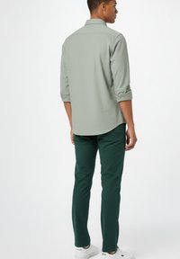 Hombre vistiendo una camisa de manga larga verde claro con los puños remangados, pantalones verde oscuro y zapatillas blancas, de pie de espaldas contra un fondo blanco.