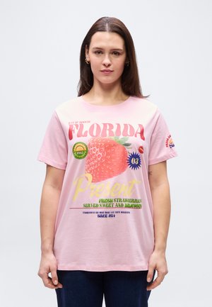 Kobieta z długimi brązowymi włosami, ubrana w różową koszulkę z dużym nadrukiem truskawki i tekstem dotyczącym Florydy oraz świeżych truskawek.