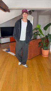 Musthave musta kapuutsiga sviiter, valge T-särk, hallid dressipüksid ja hõbedased kingad. Siseruum, kus on puitpõrand ja roheline lehtede taim.