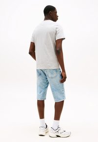 Ung mand står, iført lysegrå t-shirt, lyseblå cowboyshorts, hvide sokker og hvide sneakers på en ensfarvet hvid baggrund.