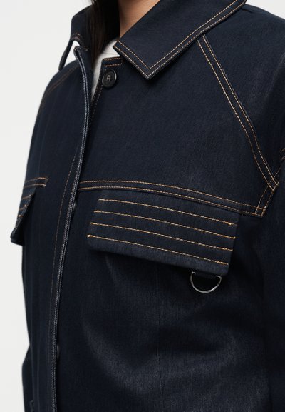 Μπλε μπουφάν με μεγάλες τσέπες με καπάκι, ραφές σε πορτοκαλί αντίθεση, κούμπωμα με κουμπιά και μικρό μεταλλικό κρίκο κάτω από το καπάκι της δεξιάς τσέπης.