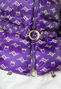 Paarse gewatteerde jas met groene logopatronen, voorzien van een gouden ronde gesp aan de taille en een ritssluiting aan de voorkant.