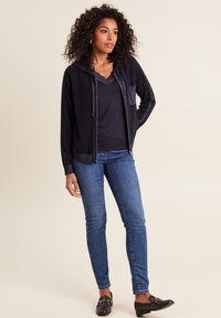 Schwarze Reißverschlusskapuzenjacke mit strukturierten Akzenten und einer Fronttasche, getragen über einem dunklen Oberteil und kombiniert mit hellblauen Skinny-Jeans und flachen Schuhen.
