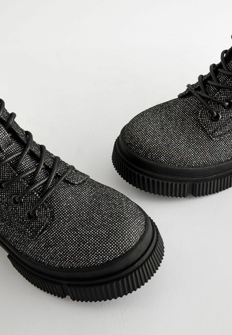 Bottes noires à lacets avec semelles épaisses en caoutchouc rainuré et un motif de surface texturé et scintillant, vues sous un angle supérieur latéral.