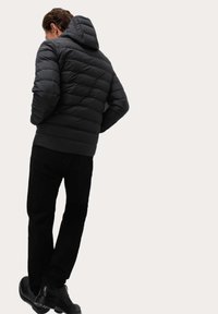 Sort pufferjakke med quiltet design, hætte og elastiske manchetter, sammen med sorte bukser og sko, set fra ryggen mod en lys baggrund.