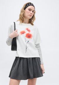 Νεαρή γυναίκα που φοράει λευκό μπλουζάκι με ανθισμένο σχέδιο, μακριά γκρι μανίκια από μέσα, γκρι φούστα με λεπτές ρίγες, μαύρη κορδέλα και κρατάει μαύρη τσάντα.