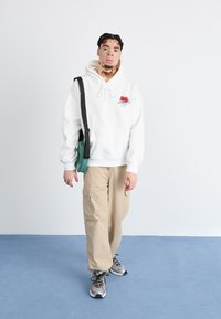 White hoodie with cherry graphic, beige cargo pants, and multi-colored sneakers. A small green bag is worn crossbody. Light background.