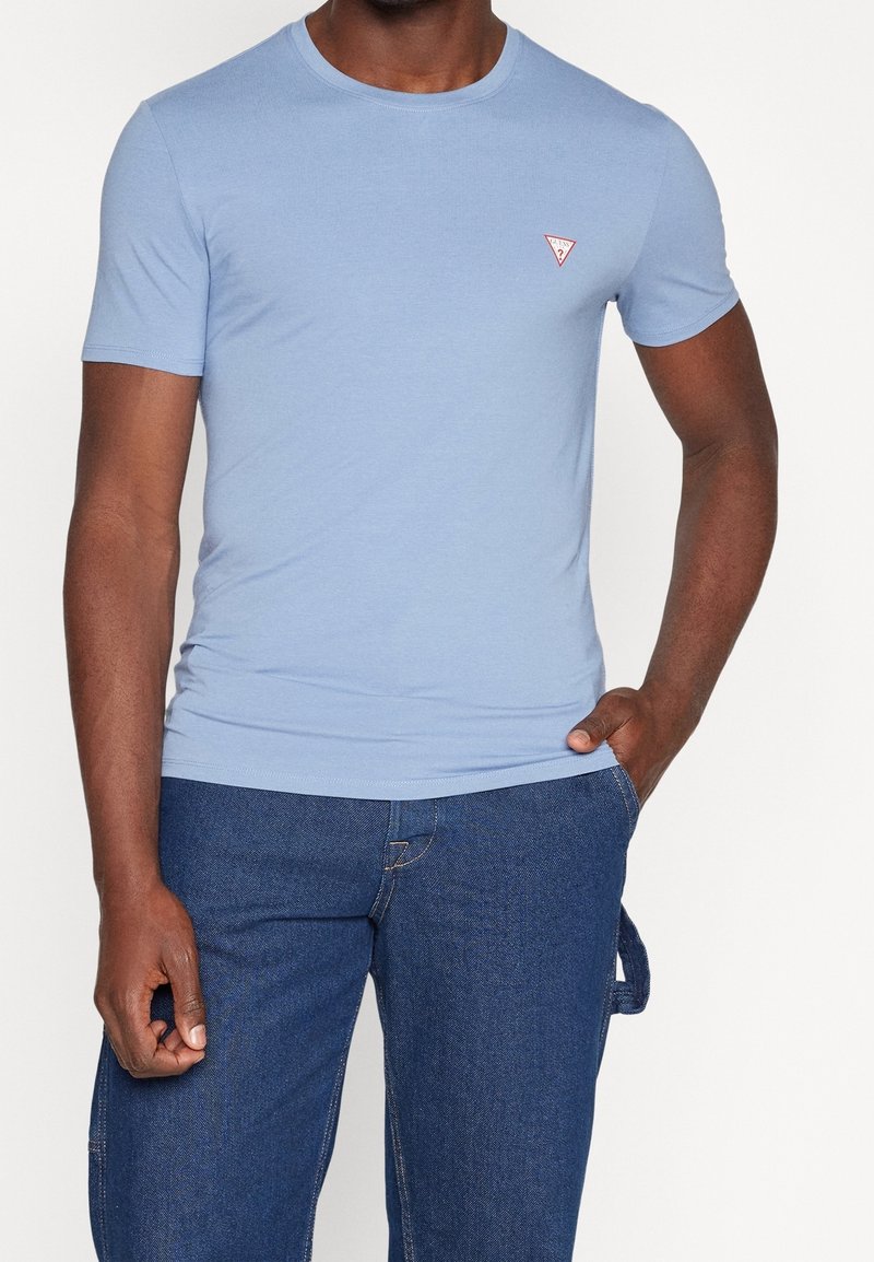 Homme portant un t-shirt bleu clair à manches courtes avec un petit logo rouge en forme de triangle sur la poitrine, assorti à un pantalon en denim bleu.