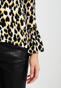 Blouse à motif léopard avec des couleurs noire, jaune et blanche. Présente des manches évasées et est associée à une jupe en cuir noir brillant.