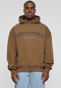 Brauner Kapuzenpullover mit dem Text "VERBOTEN", Kängurutasche, gerippten Bündchen und Saum, getragen über hellblauen Jeans.