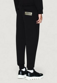 Persona vestida con pantalones de chándal negros de Armani Exchange con un logo en el bolsillo trasero, zapatillas negras con suelas blancas y calcetines blancos.