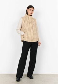 Vest bej matlasat cu guler înalt, buzunare cu fermoar și textură striată. Asortat cu un top ușor cu mânecă lungă și pantaloni negri.