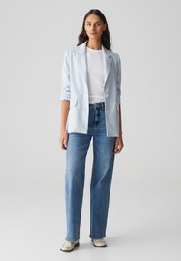 Blazer azzurro chiaro con doppi bottoni, tasche frontali, indossato sopra una maglietta bianca e jeans blu a gamba dritta. Tessuto liscio e vestibilità su misura.