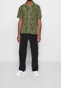 Camicia verde a maniche corte con motivo foglioso, abbinata a pantaloni neri. Scarpe da ginnastica bianche completano l'outfit.