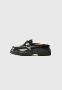 Black leather slip-on loafer with a thick black rubber sole, featuring a decorative silver strap with wing accents and stitching details.