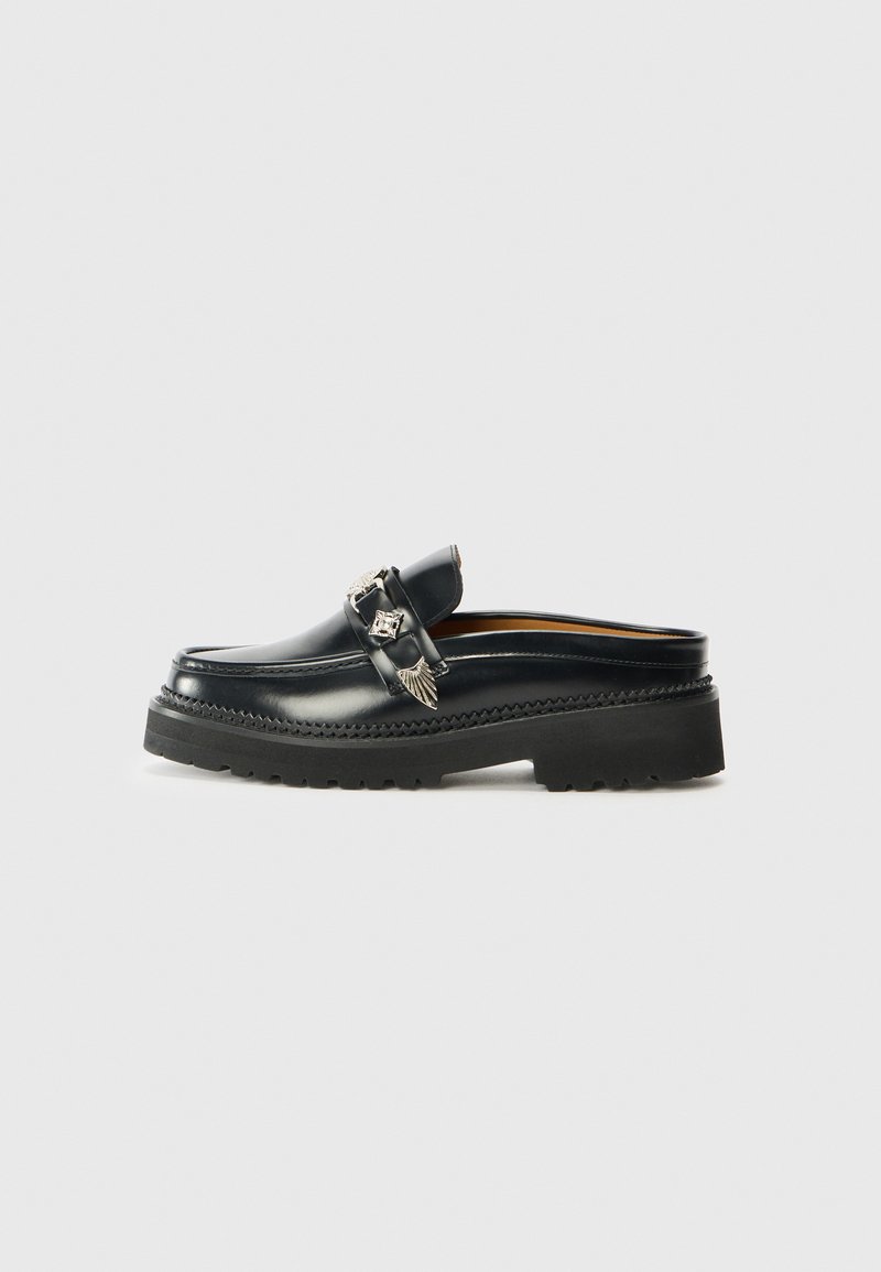 Black leather slip-on loafer with a thick black rubber sole, featuring a decorative silver strap with wing accents and stitching details.