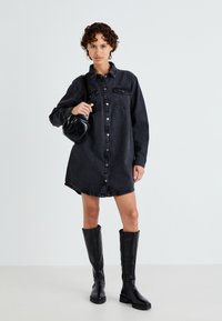 Vestido de camisa de mezclilla negra con manga larga, cierre de botones en la parte delantera, dos bolsillos en el pecho y un dobladillo curvado, combinado con botas negras hasta la rodilla.