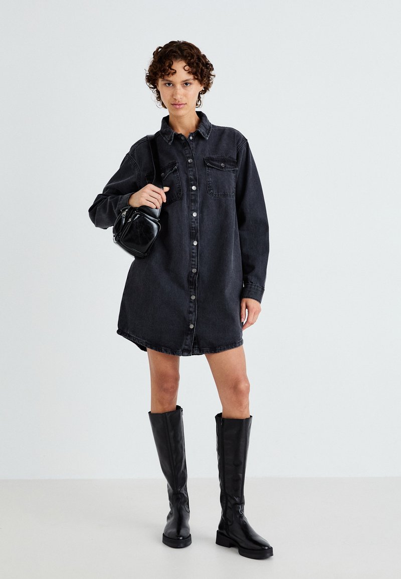Schwarzes Denim-Shirtkleid mit langen Ärmeln, Knopfverschluss vorne, zwei Brusttaschen und abgerundetem Saum, kombiniert mit knielangen schwarzen Stiefeln.