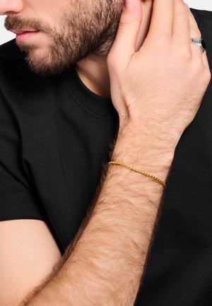 Bearded man in black shirt touching his neck, wearing a thin gold chain bracelet on his wrist and a silver ring on his finger.
