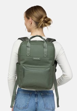 Frau mit blondem Haar zu einem Dutt gebunden, trägt ein weißes langärmeliges Oberteil und blaue Jeans, und trägt einen grünen rechteckigen Rucksack mit Frontreißverschlusstasche.