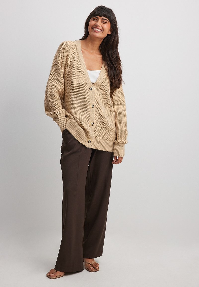 Cardigan en tricot beige avec un col en V, des boutons frontaux et un ourlet côtelé, associé à un pantalon large marron et des sandales. Texture lisse.