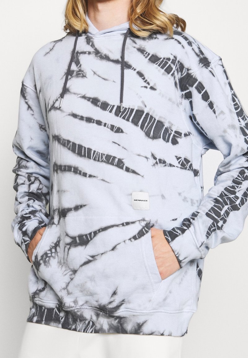 Persona con una sudadera de tie-dye en azul claro y negro, con bolsillos frontales y cordones, las manos metidas en los bolsillos, recortada por debajo de la barba.