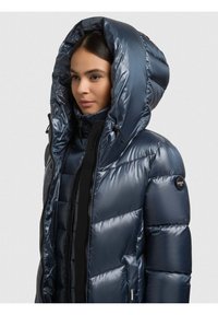 Tamsiai mėlyna pūkinė striukė su aukšta apykakle ir dideliu gobtuvu. Yra kišenės su užtrauktukais, blizgus paviršius ir prekės ženklo logotipas ant rankovės.