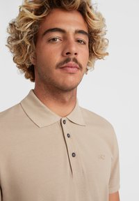 Beige polo skjorte med en tekstureret finish, tre mørke knapper og et lille broderet logo på venstre bryst. Krave-design.