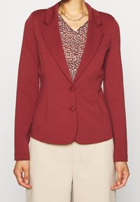 Blazer marron avec une coupe ajustée, deux boutons et des revers pointus. Porté sur un haut à motif avec un décolleté en V et des couleurs subtiles.