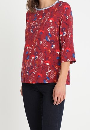 Top rosso a fantasie con design floreale blu e bianco, maniche corte e scollatura rotonda con rifinitura a contrasto. Indossato con jeans scuri.