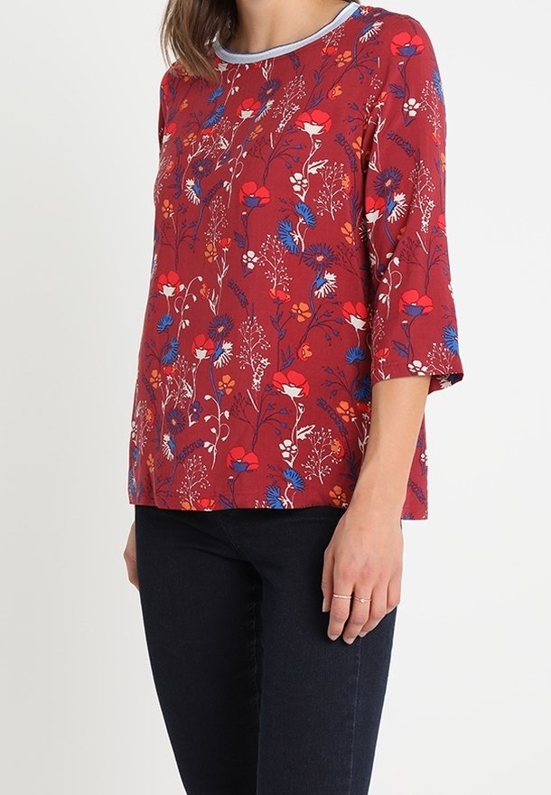 Top rosso a fantasie con design floreale blu e bianco, maniche corte e scollatura rotonda con rifinitura a contrasto. Indossato con jeans scuri.