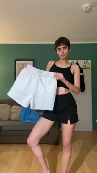 Weiße gefaltete Shorts mit elastischem Bund und Kordelzug. Schwarzes Oberteil daneben, vor einer grünen Wand und einem Holzfußboden.