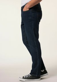 Jeans in denim blu scuro con taglio dritto, cinque tasche e dettagli di cucitura gialla, abbinati a sneakers nere e bianche.