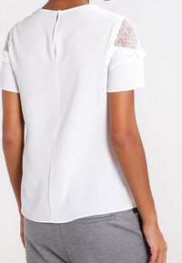 Chemisier blanc à manches courtes avec accents en dentelle, bord froncé et dos en forme de clé. Texture lisse avec un subtil éclat.