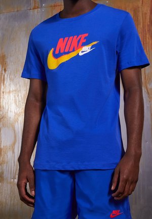 Osoba ubrana w niebieski strój Nike z czerwonymi i żółtymi logo Nike na koszulce oraz czerwonym logo na szortach na tle zardzewiałego metalu.