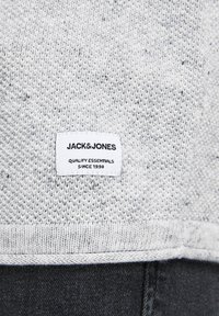 Tessuto di maglione lavorato a maglia grigio chiaro con una superficie testurizzata. Presenta un'etichetta bianca con testo nero: "JACK & JONES QUALITY ESSENTIALS SINCE 1990."