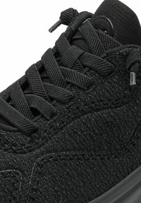 Black sneaker with a textured woven upper, flat laces, and a rubber sole. Features include subtle stitching and a loop detail on the tongue.