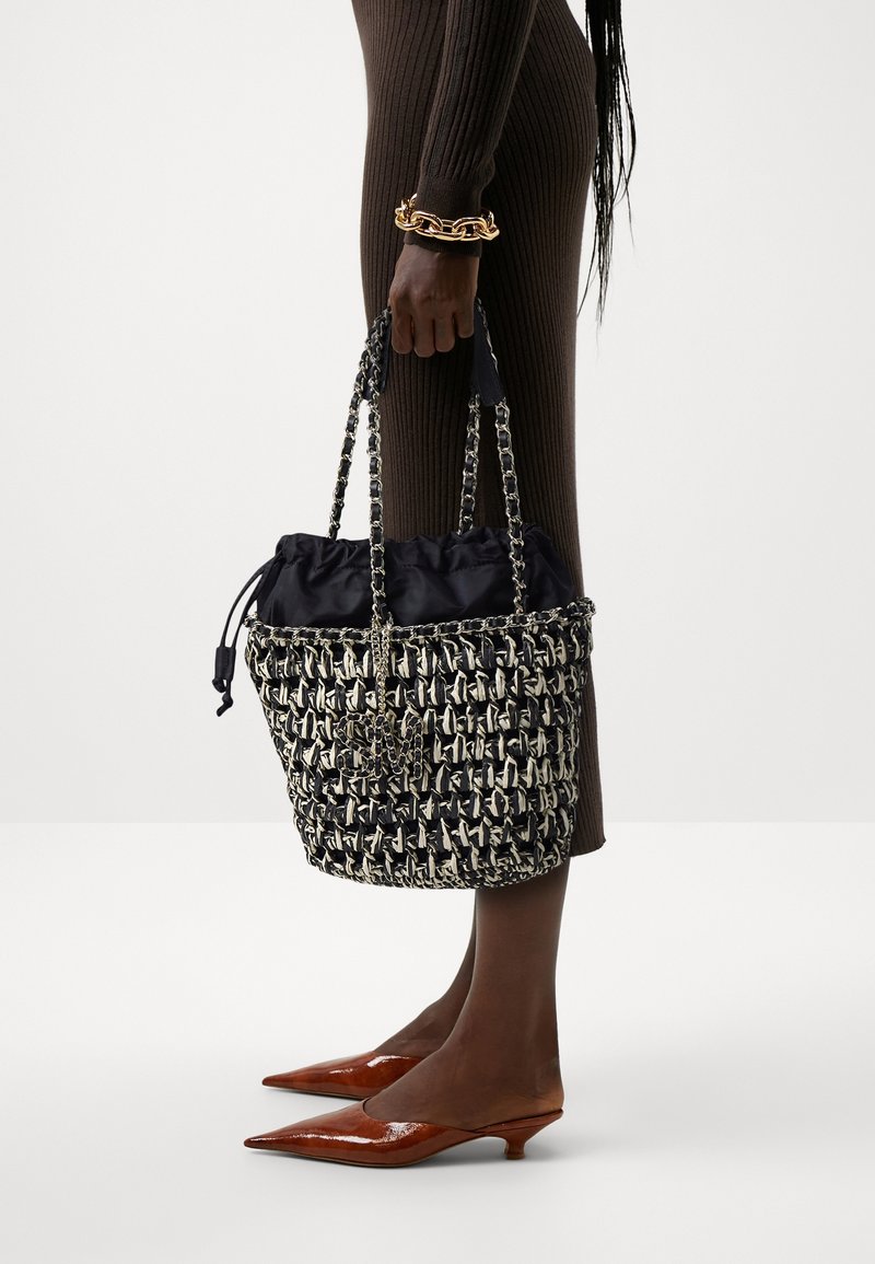 Woven black and white bucket bag with a drawstring closure, chain handles, and a soft black lining; structured shape and textured design.