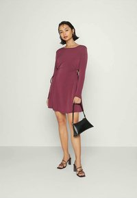 Mulher vestindo um vestido longo de cor ameixa com recortes laterais, segurando uma bolsa preta e usando sandálias de salto com tiras, em um fundo simples.