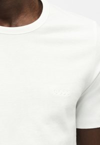 Gros plan d'une personne à la peau foncée portant un t-shirt blanc uni avec un petit logo BOSS en relief sur la poitrine.