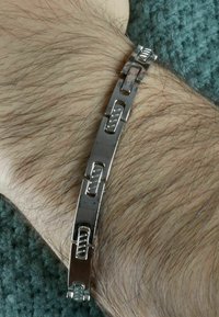 Bracciale in metallo con maglie intrecciate, superficie lucida, forme rettangolari e accenti testurizzati sulle maglie. Posizionato su un polso.