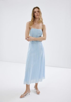 Mujer con un vestido midi sin mangas azul claro y sandalias blancas de tacón, de pie con los brazos cruzados frente a un fondo blanco liso.